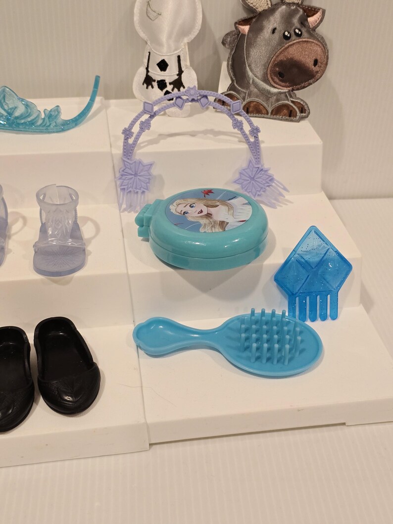K&ouml;nnte beinhalten: Eine Sammlung von Kinderspielzeug und Zubeh&ouml;r, darunter eine blaue Haarb&uuml;rste, schwarze Schuhe und ein blauer Kompaktpuder mit einem Charakterbild. Weitere Artikel sind ein Diadem, ein klarer Schuh und ein blaues, eisgeformtes Accessoire.