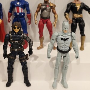 May include: A collection of action figures featuring various superheroes. The figures include Captain America in a blue and red suit, a figure in a black and gray suit, and a gray Batman figure with a black bat symbol.
