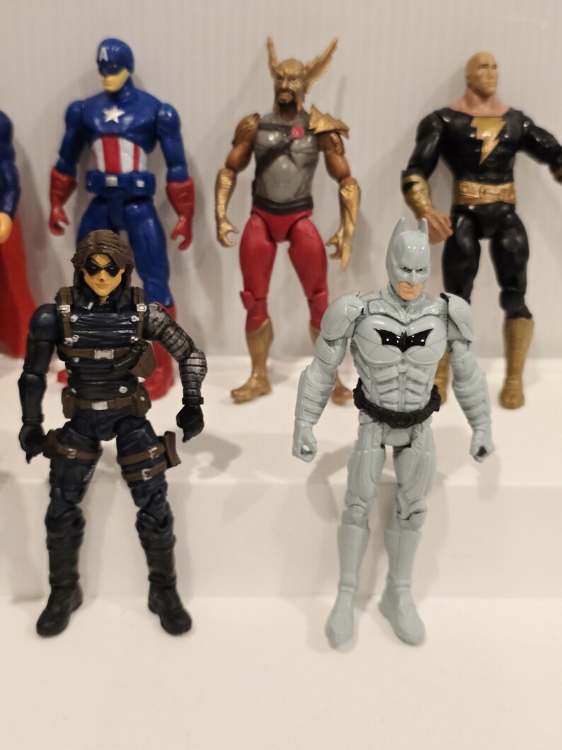 May include: A collection of action figures featuring various superheroes. The figures include Captain America in a blue and red suit, a figure in a black and gray suit, and a gray Batman figure with a black bat symbol.