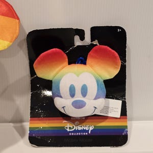 May include: A Mickey Mouse plush toy with rainbow-colored ears and face, attached to a black cardboard backing. The packaging features the Disney Collection logo and the word "Rainbow". The toy is designed for ages 2+.