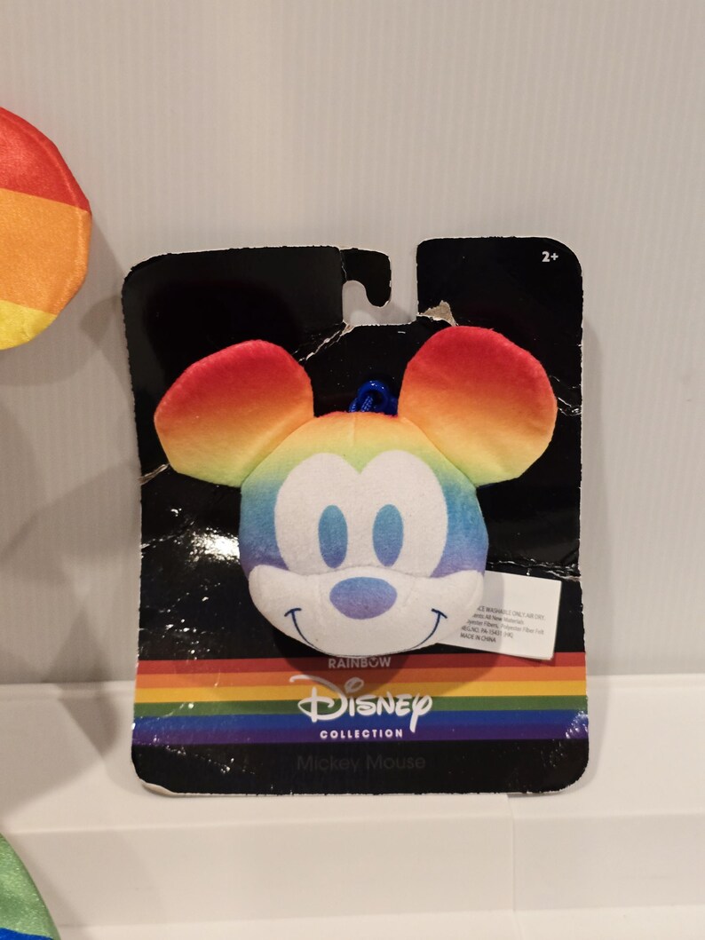 May include: A Mickey Mouse plush toy with rainbow-colored ears and face, attached to a black cardboard backing. The packaging features the Disney Collection logo and the word "Rainbow". The toy is designed for ages 2+.