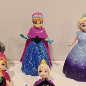 May include: A selection of small plastic figurines depicting characters from the animated film. One figurine is in a blue dress with glitter, a pink cape, and a matching hat. Other figurines are also visible.