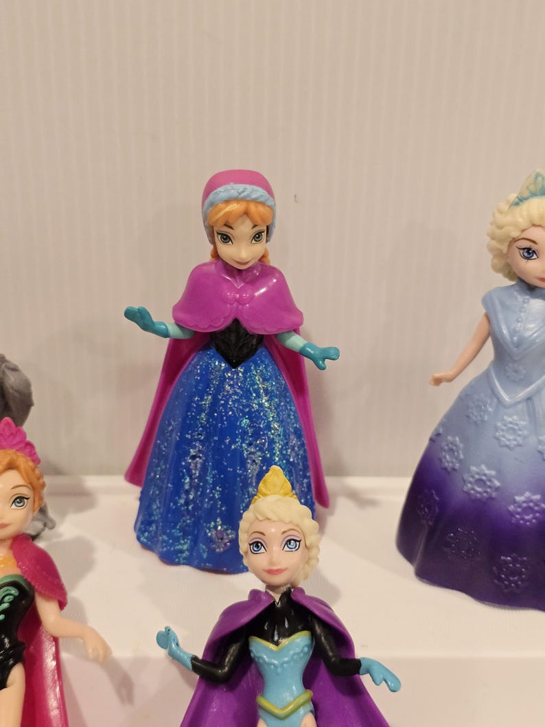 May include: A selection of small plastic figurines depicting characters from the animated film. One figurine is in a blue dress with glitter, a pink cape, and a matching hat. Other figurines are also visible.