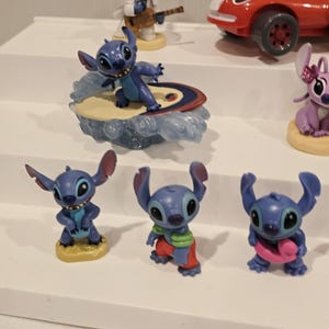 May include: A collection of blue Stitch figurines in various poses. One figurine surfs on a surfboard, while another plays a guitar. Other figurines are in different outfits, including a red race car.
