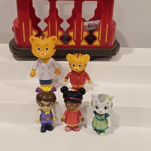 May include: A red toy trolley with yellow accents, and six character figurines. The figurines include a tiger, a cat, and other characters wearing various outfits. The toys are made of plastic and are designed for children.