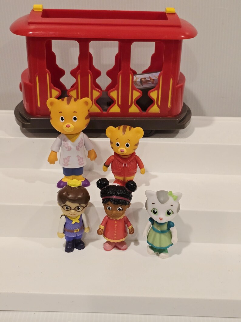 May include: A red toy trolley with yellow accents, and six character figurines. The figurines include a tiger, a cat, and other characters wearing various outfits. The toys are made of plastic and are designed for children.