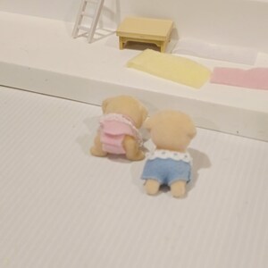 Puede incluir: Dos figuras de animales de juguete en miniatura, una con un atuendo rosa y la otra con pantalones cortos azules con ribete blanco. Las figuras son de color marr&oacute;n claro y parecen ser parte de un juego. Otros accesorios en miniatura son visibles.