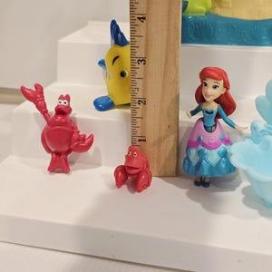 May include: A collection of colorful figurines featuring characters from a popular animated film. The figures include a red crab, a yellow fish with blue fins, and a doll-like figure with red hair and a blue and pink dress. A wooden ruler is next to the figurines.