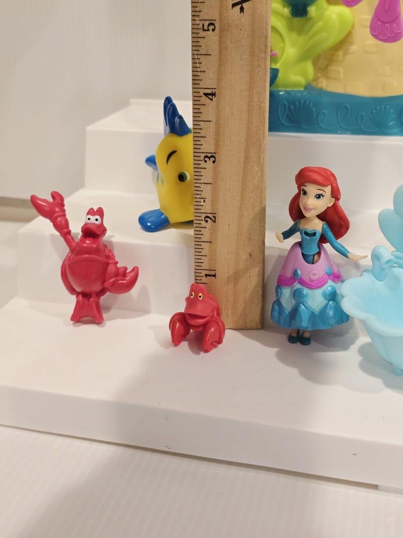 May include: A collection of colorful figurines featuring characters from a popular animated film. The figures include a red crab, a yellow fish with blue fins, and a doll-like figure with red hair and a blue and pink dress. A wooden ruler is next to the figurines.