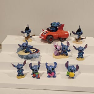 May include: A collection of blue Stitch figurines in various poses. Some figurines are playing a guitar, surfing, or driving a red car. Other figurines are holding a pineapple or wearing accessories.