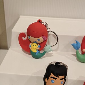 May include: Ariel keychain featuring a red-haired mermaid holding a yellow and green fish. The keychain has a silver metal ring. Other character keychains are visible in the background.