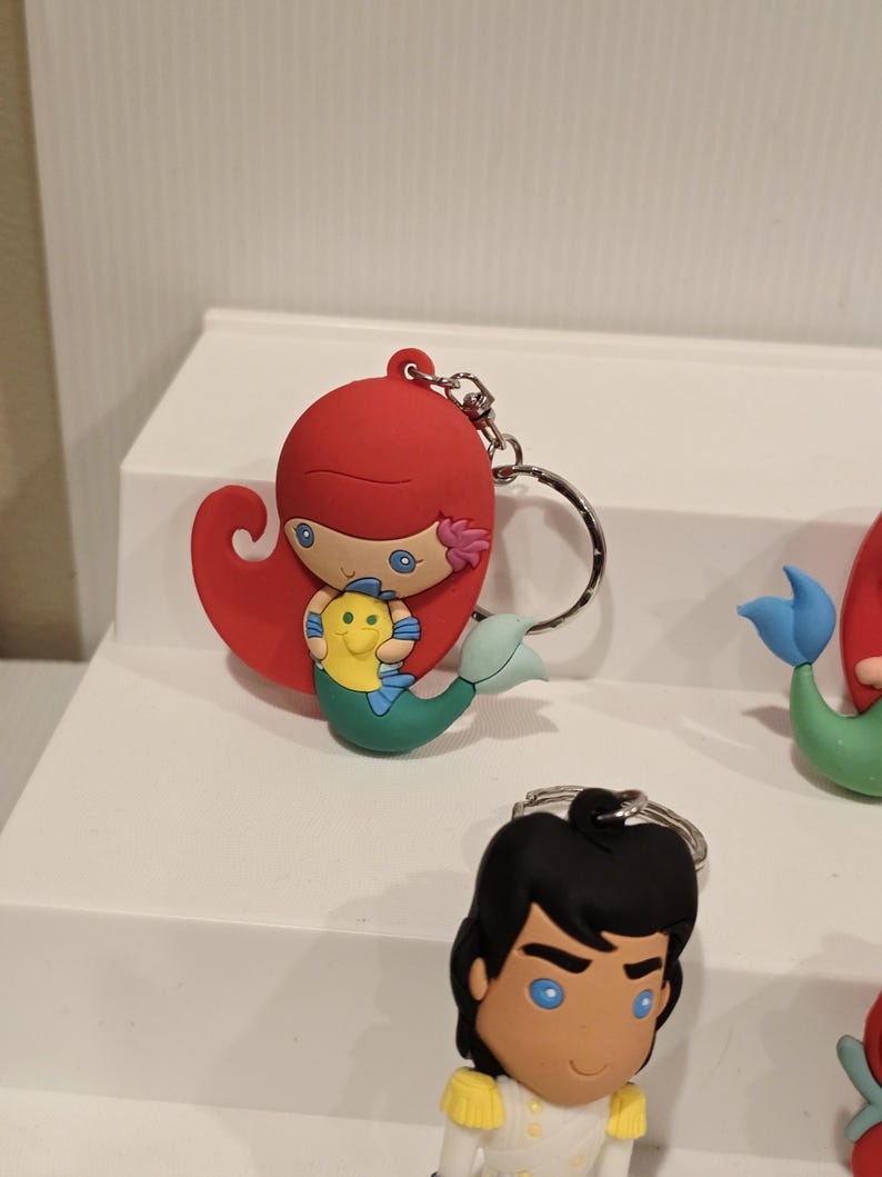 May include: Ariel keychain featuring a red-haired mermaid holding a yellow and green fish. The keychain has a silver metal ring. Other character keychains are visible in the background.