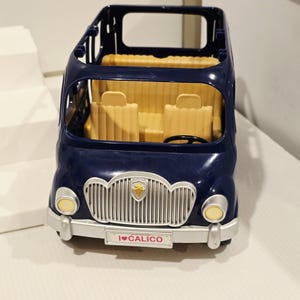 May include: A navy blue toy bus with a silver grill and yellow headlights. The interior features yellow seats and a steering wheel. The front of the bus has a license plate that reads "I ❤️ CALICO".