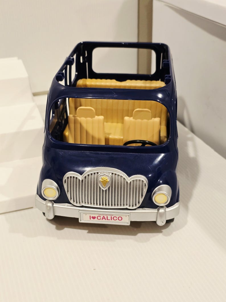 May include: A navy blue toy bus with a silver grill and yellow headlights. The interior features yellow seats and a steering wheel. The front of the bus has a license plate that reads "I ❤️ CALICO".