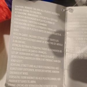 May include: A gray tag with text in multiple languages, including English, Italian, and Spanish. The text warns about removing tags and plastic fasteners before giving the item to children. The tag is attached to a colorful, rainbow-colored fabric.