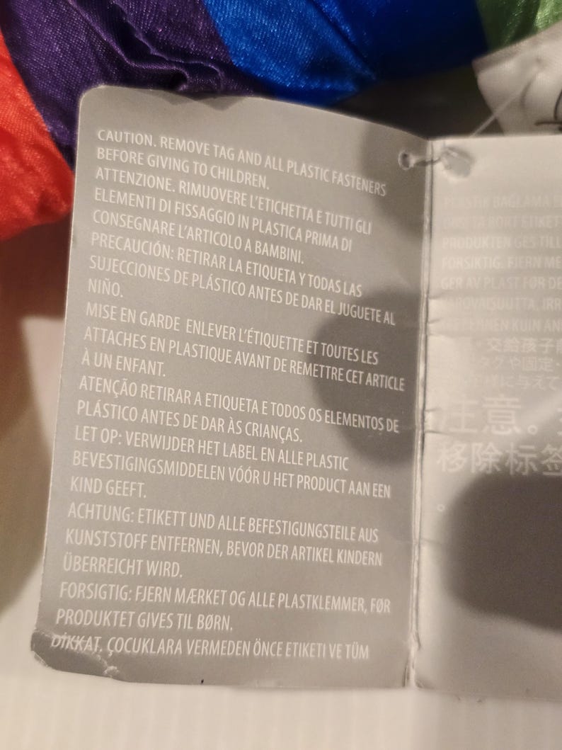May include: A gray tag with text in multiple languages, including English, Italian, and Spanish. The text warns about removing tags and plastic fasteners before giving the item to children. The tag is attached to a colorful, rainbow-colored fabric.
