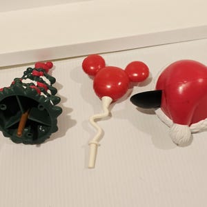 May include: Three plastic holiday-themed ornaments: a dark green Christmas tree with white snow accents, a red balloon-shaped icon with three circular ears, and a red Santa hat with black ears and a white pom-pom trim on a white surface.