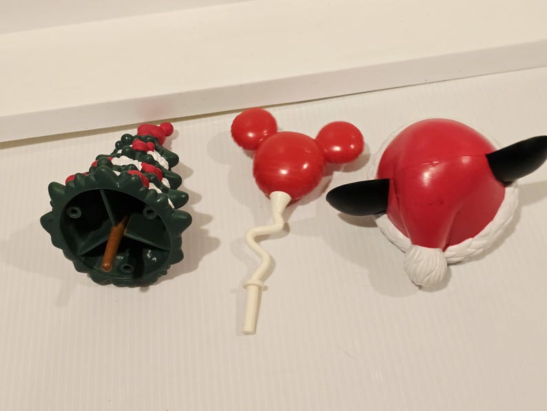 May include: Three plastic holiday-themed ornaments: a dark green Christmas tree with white snow accents, a red balloon-shaped icon with three circular ears, and a red Santa hat with black ears and a white pom-pom trim on a white surface.