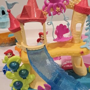 K&ouml;nnte beinhalten: Ariel's Secret Spielset mit einem gelben und rosa Schloss mit einer blauen Rutsche, einem rosa Oktopus und einer rosa Muschelschaukel. Das Set enth&auml;lt eine kleine Ariel-Figur. F&uuml;r Kinder konzipiert.