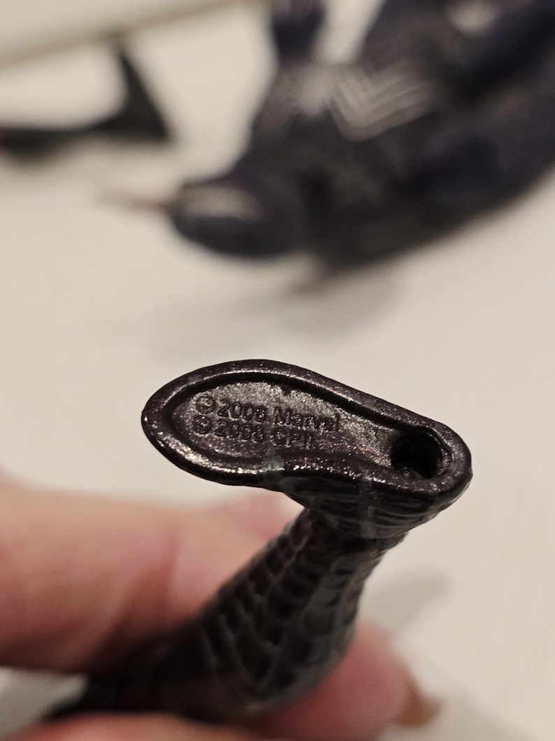 May include: Close-up of a dark, textured, snake-like figurine. The base of the figurine has the text "&copy;2006 Marvel" and "&copy;2006 CPI". The figurine is dark in color and appears to be made of plastic.