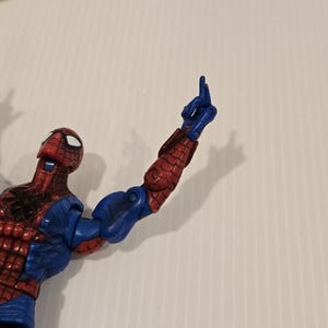 Pode incluir: Uma figura de a&ccedil;&atilde;o do Homem-Aranha, predominantemente vermelha e azul, com um design texturizado detalhado. O bra&ccedil;o da figura est&aacute; levantado, com o dedo indicador apontando para cima. A figura est&aacute; sobre um fundo branco liso.