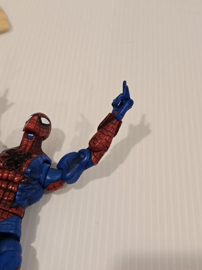 Pode incluir: Uma figura de a&ccedil;&atilde;o do Homem-Aranha, predominantemente vermelha e azul, com um design texturizado detalhado. O bra&ccedil;o da figura est&aacute; levantado, com o dedo indicador apontando para cima. A figura est&aacute; sobre um fundo branco liso.