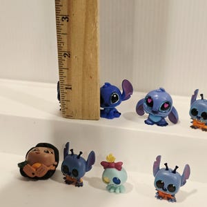 May include: A collection of small, colorful figurines featuring characters from the animated film Lilo & Stitch. The figurines are primarily blue, with some orange and pink accents. A wooden ruler is placed next to the figurines.