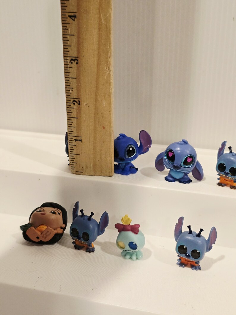 May include: A collection of small, colorful figurines featuring characters from the animated film Lilo & Stitch. The figurines are primarily blue, with some orange and pink accents. A wooden ruler is placed next to the figurines.
