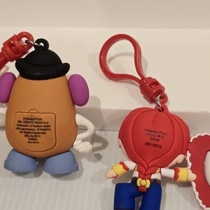 May include: Two Disney Pixar character keyrings. One is Mr. Potato Head, tan with a black hat, purple ears, and blue feet. The other is Jessie, with a red hat, blonde braid, blue trousers, and yellow sleeves. Both have red key rings.