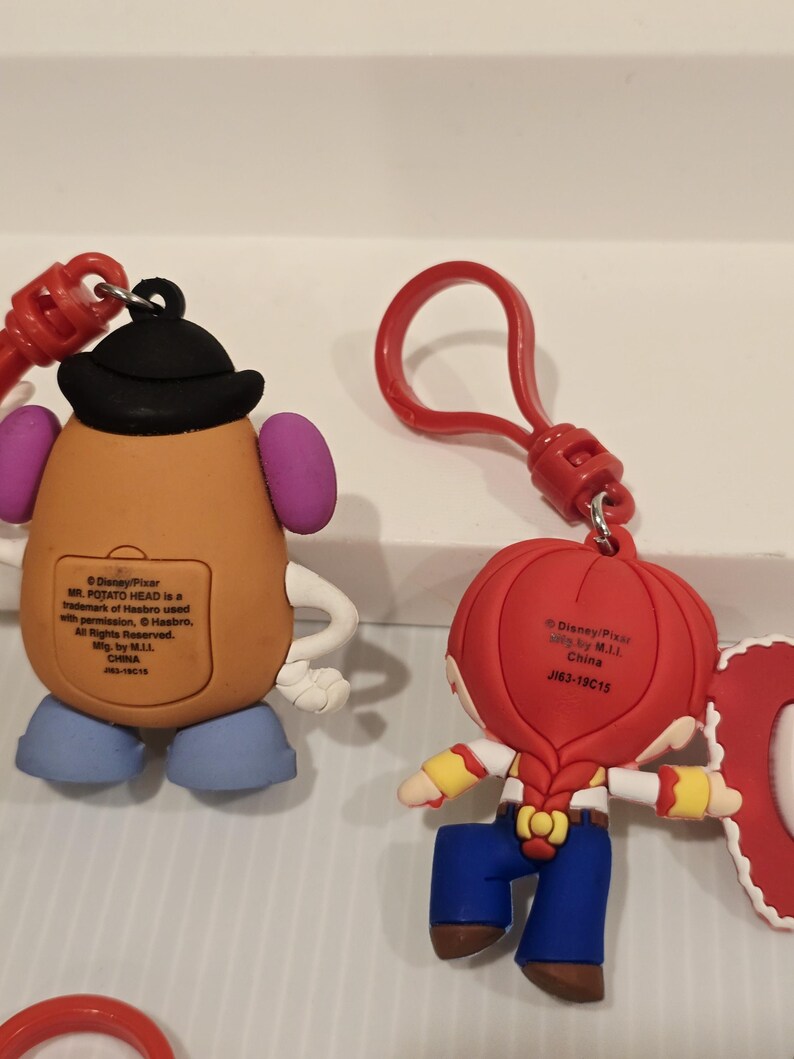May include: Two Disney Pixar character keyrings. One is Mr. Potato Head, tan with a black hat, purple ears, and blue feet. The other is Jessie, with a red hat, blonde braid, blue trousers, and yellow sleeves. Both have red key rings.