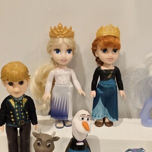 May include: A collection of Disney Frozen character dolls, including Elsa and Anna, each wearing a golden crown. Other characters include Kristoff, Olaf, and a reindeer. The dolls are on a white shelf.