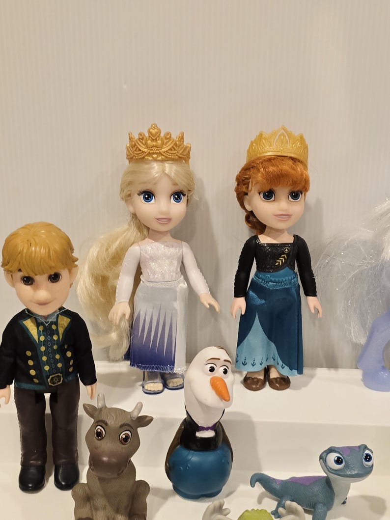 May include: A collection of Disney Frozen character dolls, including Elsa and Anna, each wearing a golden crown. Other characters include Kristoff, Olaf, and a reindeer. The dolls are on a white shelf.