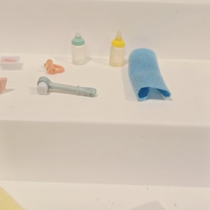 Puede incluir: Colecci&oacute;n de accesorios de beb&eacute; en miniatura, que incluye dos biberones, un cepillo de dientes de dedo azul, un chupete y un pa&ntilde;o azul. Los art&iacute;culos est&aacute;n dispuestos sobre una superficie blanca, lo que sugiere un entorno de guarder&iacute;a o tem&aacute;tico de beb&eacute;s.
