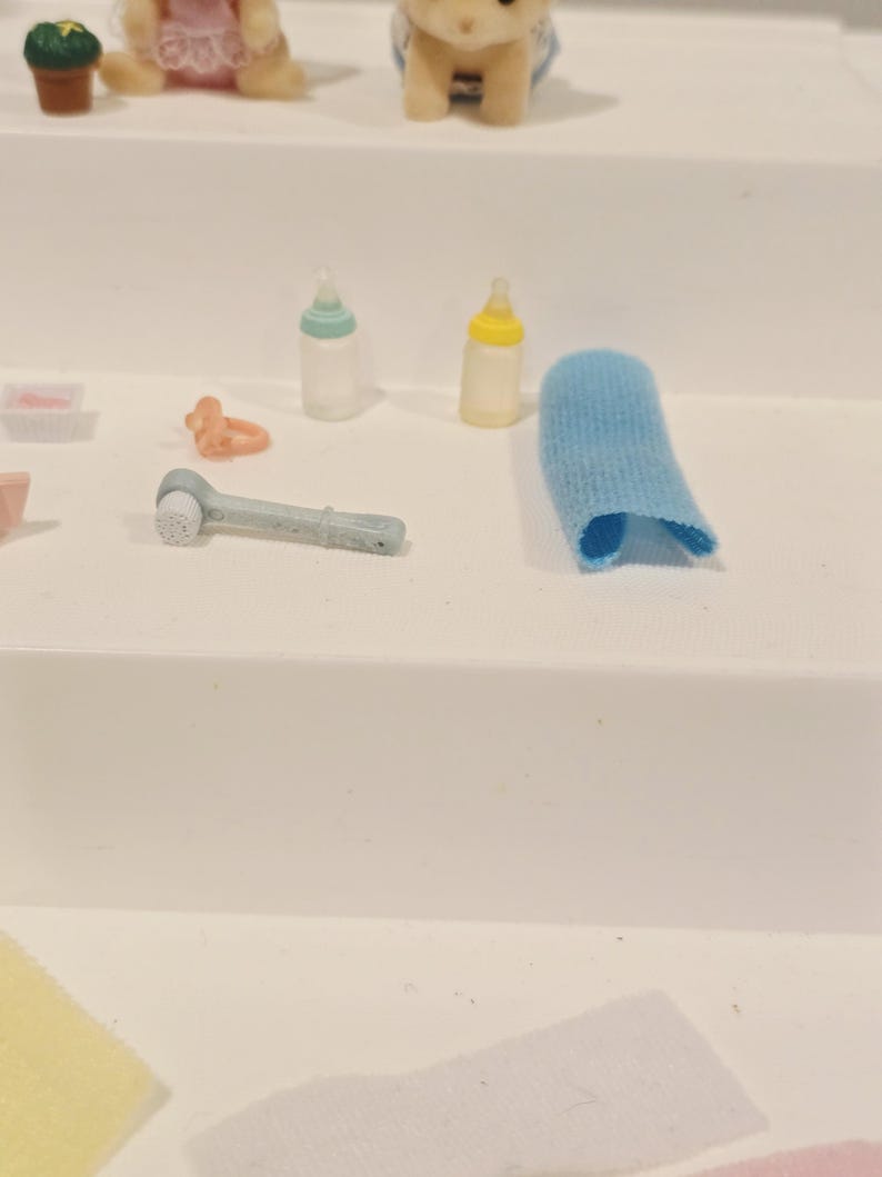 Puede incluir: Colecci&oacute;n de accesorios de beb&eacute; en miniatura, que incluye dos biberones, un cepillo de dientes de dedo azul, un chupete y un pa&ntilde;o azul. Los art&iacute;culos est&aacute;n dispuestos sobre una superficie blanca, lo que sugiere un entorno de guarder&iacute;a o tem&aacute;tico de beb&eacute;s.