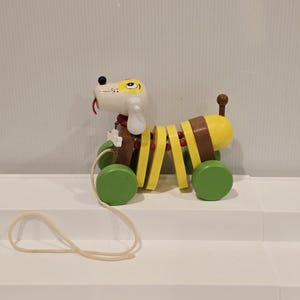 May include: A wooden pull-along toy dog with a yellow and brown segmented body, green wheels, and a white pull string. The dog has a white head with black eyes, a red tongue, and a brown tail.