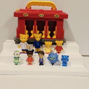 K&ouml;nnte beinhalten: Ein roter Spielzeug-Zugkoffer mit gelbem Griff, begleitet von elf Charakterfiguren. Die Figuren umfassen Tigercharaktere in verschiedenen Outfits, eine Katze, ein M&auml;dchen in einem roten Kleid, einen Jungen mit Brille, eine Eule und einen blauen Tiger.