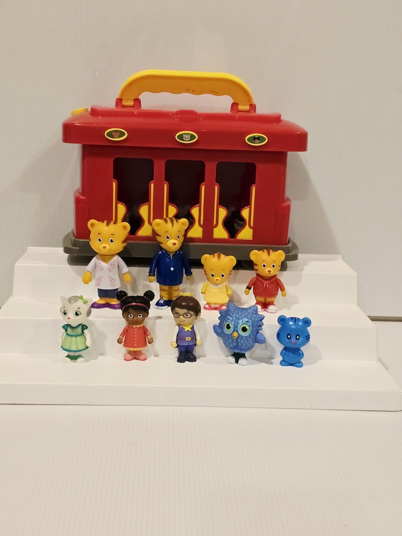 K&ouml;nnte beinhalten: Ein roter Spielzeug-Zugkoffer mit gelbem Griff, begleitet von elf Charakterfiguren. Die Figuren umfassen Tigercharaktere in verschiedenen Outfits, eine Katze, ein M&auml;dchen in einem roten Kleid, einen Jungen mit Brille, eine Eule und einen blauen Tiger.