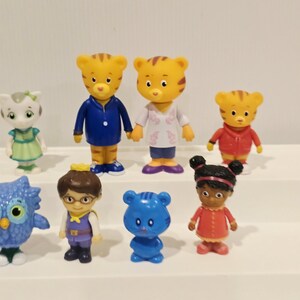May include: A collection of eight small plastic character figurines including two tiger figures, a white cat figure, a blue owl, a human figure with glasses, a small blue bear, a girl figure with dark hair, and a small tiger in a red jacket.