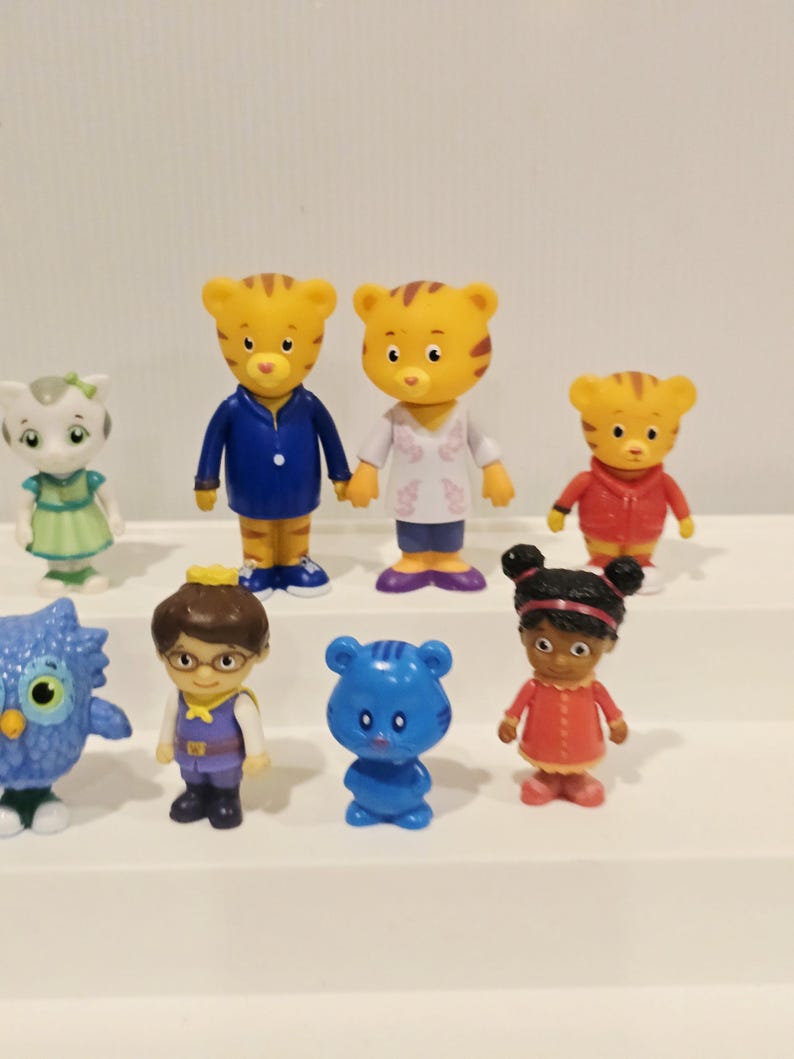 May include: A collection of eight small plastic character figurines including two tiger figures, a white cat figure, a blue owl, a human figure with glasses, a small blue bear, a girl figure with dark hair, and a small tiger in a red jacket.