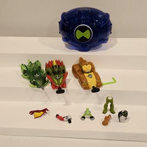 May include: A collection of Ben 10 action figures and accessories. The set includes a large blue Omnitrix, three vehicle figures in green, red, and brown, and several small character pieces. The toys are arranged on a white surface.