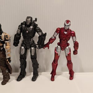 May include: Three action figures are displayed. The figure on the right is red and silver, resembling Iron Man. The middle figure is black and silver, and the figure on the left is brown and tan.