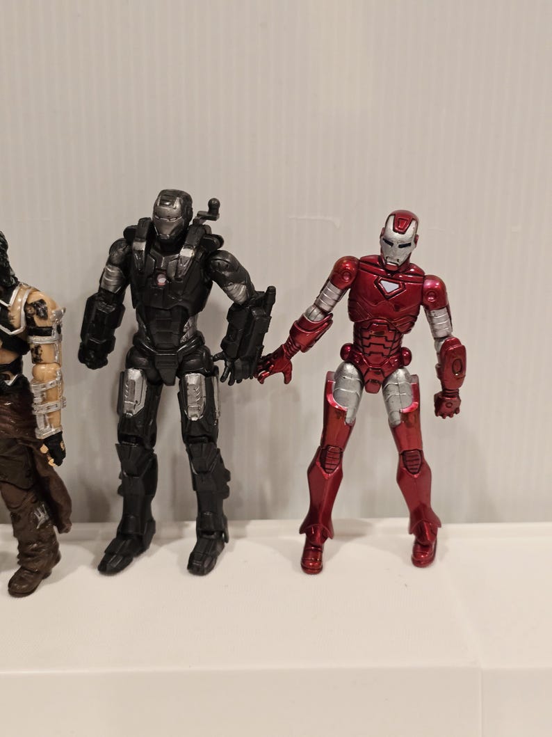 May include: Three action figures are displayed. The figure on the right is red and silver, resembling Iron Man. The middle figure is black and silver, and the figure on the left is brown and tan.