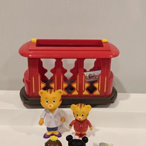 May include: A red toy trolley with yellow accents and a brown base. Several small figurines of cartoon characters are in front of the trolley. The figurines include a tiger, a cat, and other characters wearing various outfits.