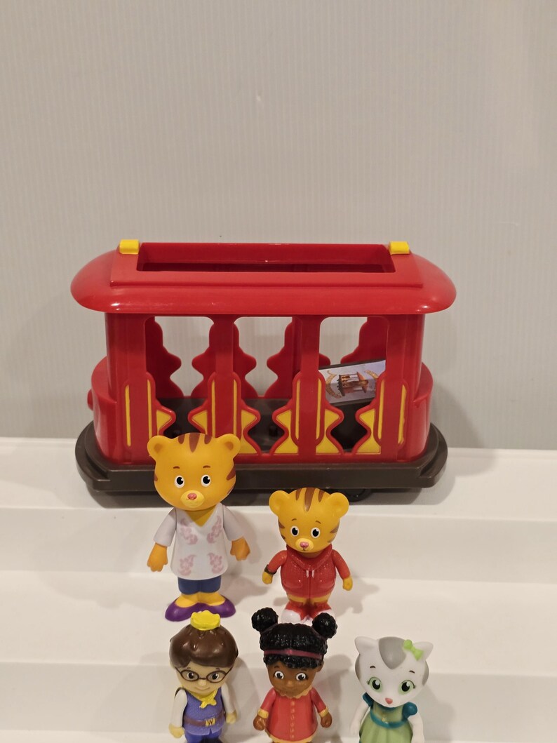 May include: A red toy trolley with yellow accents and a brown base. Several small figurines of cartoon characters are in front of the trolley. The figurines include a tiger, a cat, and other characters wearing various outfits.