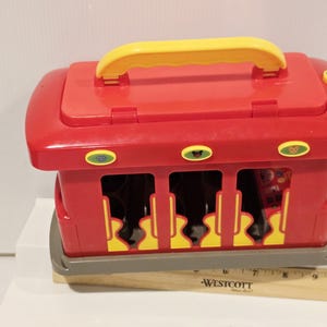 K&ouml;nnte beinhalten: Ein roter Spielzeugzug aus Kunststoff mit gelbem Griff und Akzenten. Das Spielzeug hat eine rechteckige Form mit Aussparungen an den Seiten und eine graue Basis. Der Zug hat drei ovale Kn&ouml;pfe mit Tiergrafiken.