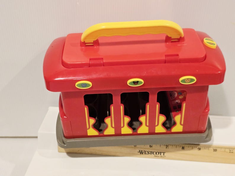 K&ouml;nnte beinhalten: Ein roter Spielzeugzug aus Kunststoff mit gelbem Griff und Akzenten. Das Spielzeug hat eine rechteckige Form mit Aussparungen an den Seiten und eine graue Basis. Der Zug hat drei ovale Kn&ouml;pfe mit Tiergrafiken.