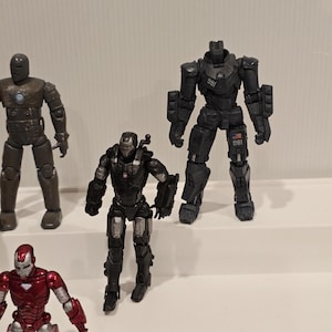 May include: Several action figures of Iron Man characters are displayed. The figures are in various poses and colors, including red, silver, black, and brown. The figures are on a white surface.