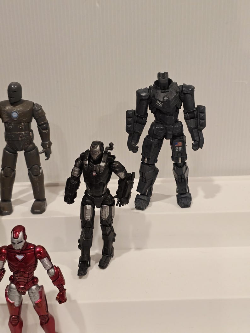 May include: Several action figures of Iron Man characters are displayed. The figures are in various poses and colors, including red, silver, black, and brown. The figures are on a white surface.