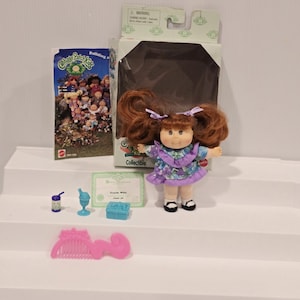 May include: A Cabbage Patch Kids collectible doll with red hair in pigtails, wearing a purple floral dress and black shoes. Accessories include a comb, a certificate, and miniature toys. The doll is in its original packaging.