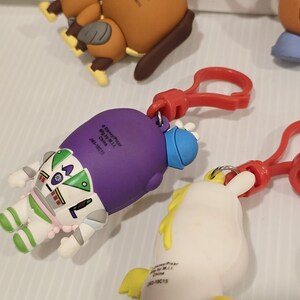 May include: A selection of character keyrings, featuring a brown dog, Buzz Lightyear, and a white unicorn. Each keyring has a red clasp. The keyrings are made from a rubbery material and are brightly coloured.